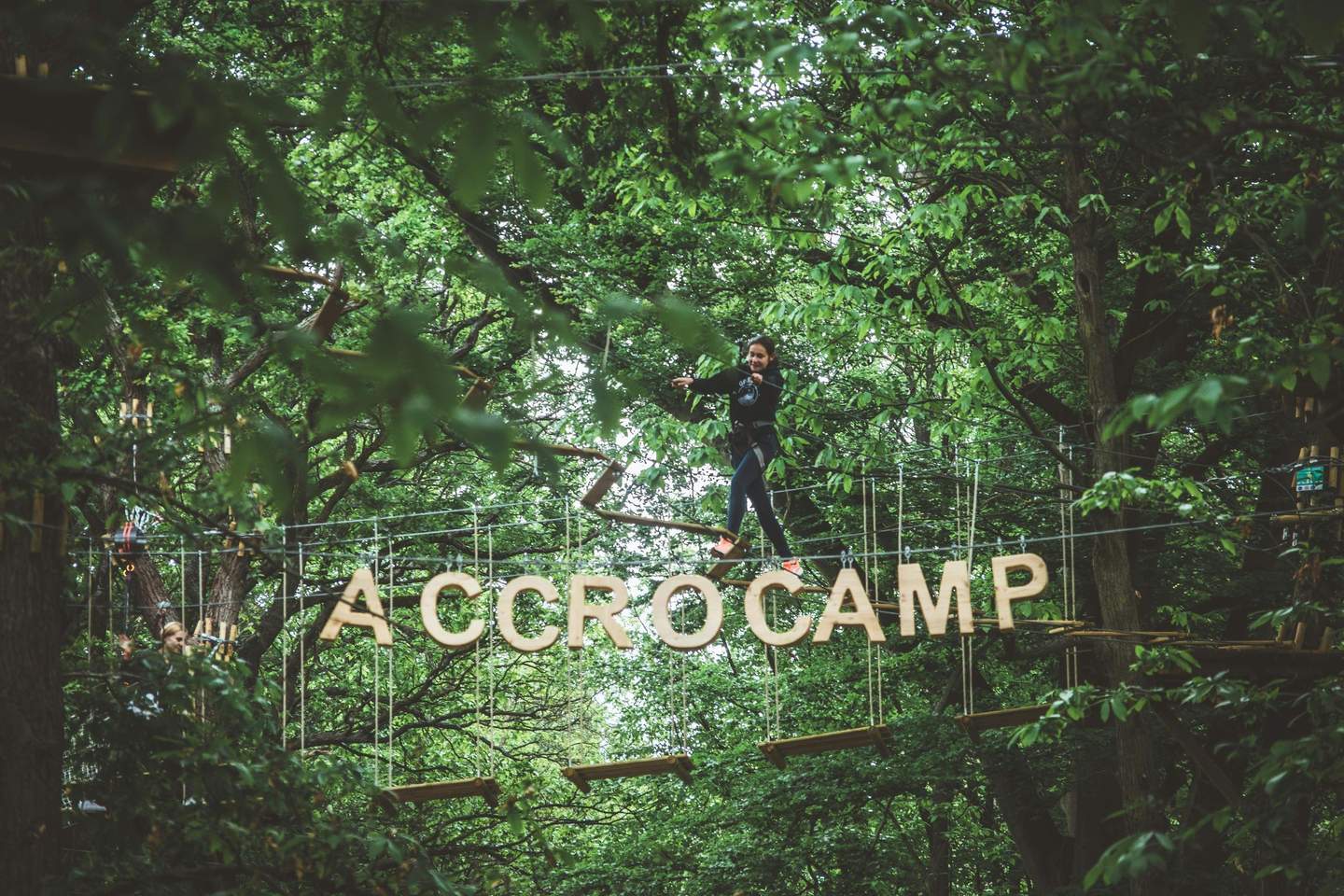 AccroCamp: Paris-Jablines Eine Person balanciert auf einem Hochseilgarten in einem Wald, unter ihr stehen große Holzbuchstaben mit der Aufschrift "ACCROCAMP".