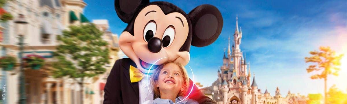 A child smiles up at a person in a Mickey Mouse costume, with a fairytale castle and colorful buildings in the background.