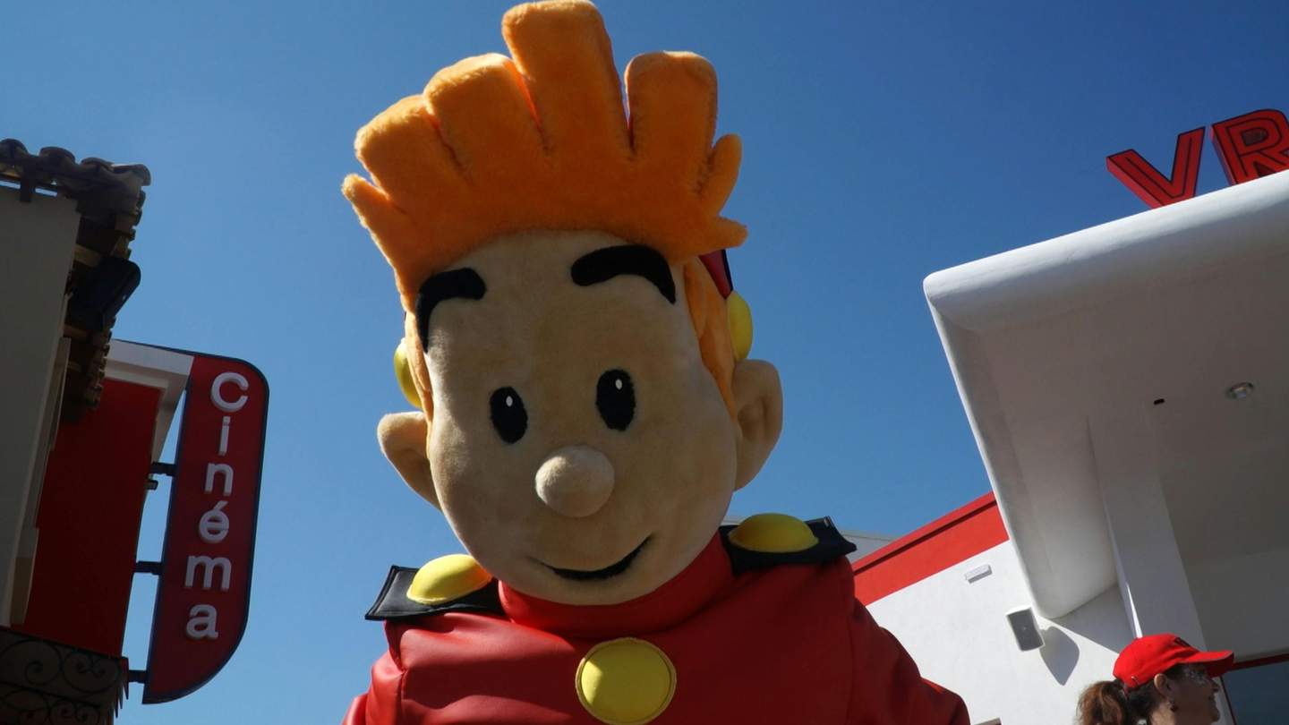 Large mascot with orange hair in a red outfit stands outside, next to a building with "Cinéma" sign and another white structure.