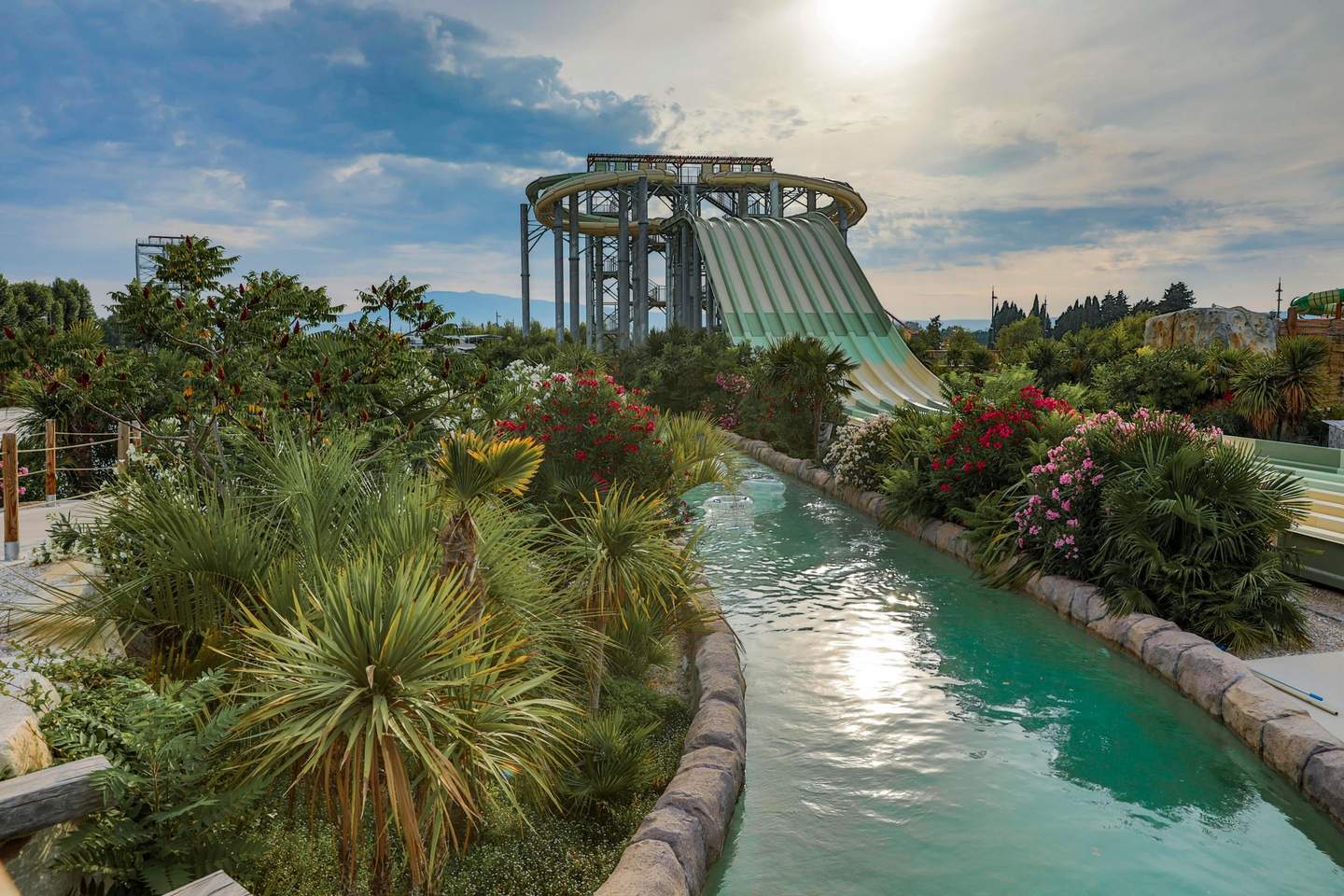 Una struttura di scivoli d'acqua e un lazy river delimitati da piante verdi e fiori vivaci sotto un cielo nuvoloso.
