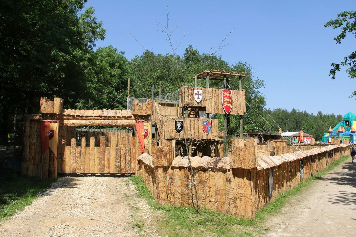 Wooden castle structure with towers, walkways, and flag banners, set in a forested area under a clear blue sky.