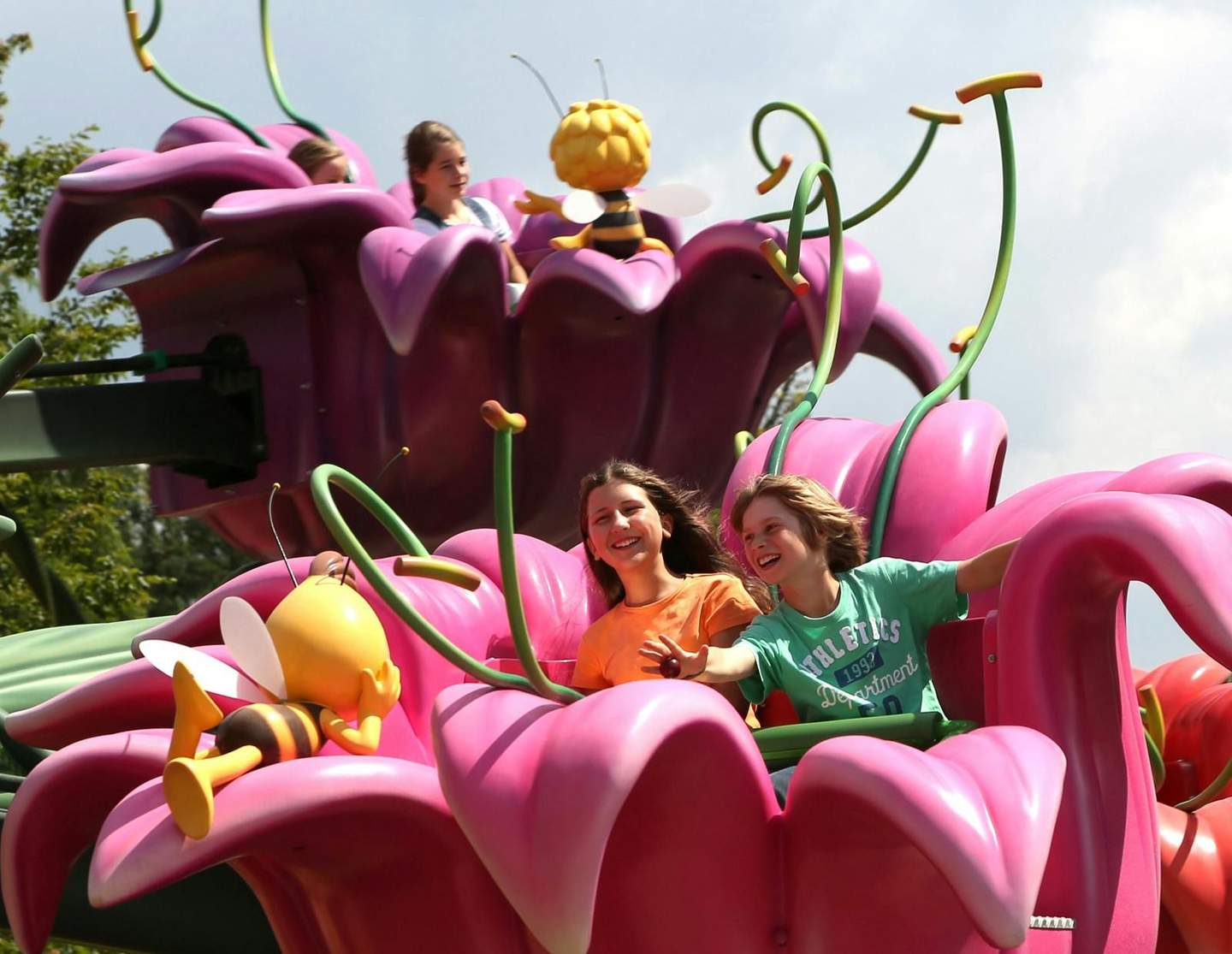 Two children smiling on a pink flower-shaped amusement ride with bee figures, set against a background of trees and sky.