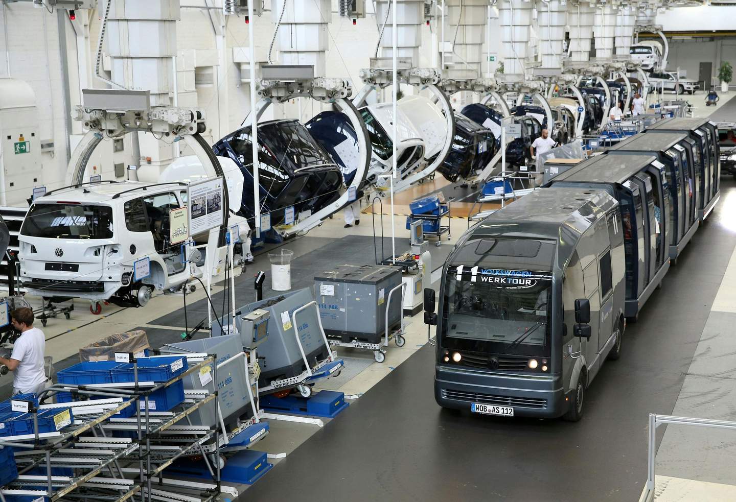An automotive assembly line with partially assembled car bodies and a tour vehicle labeled "Volkswagen WerkTour" driving through.