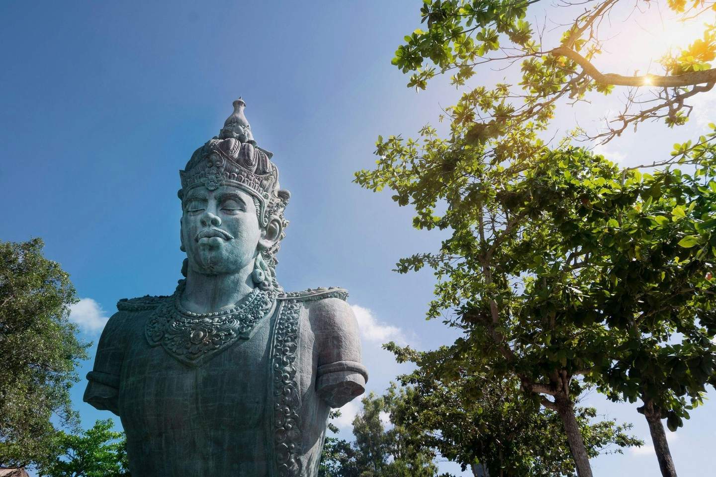 Garuda Wisnu Kencana Cultural Park A large stone statue of a crowned figure, surrounded by green trees under a bright blue sky with sunlight filtering through branches.