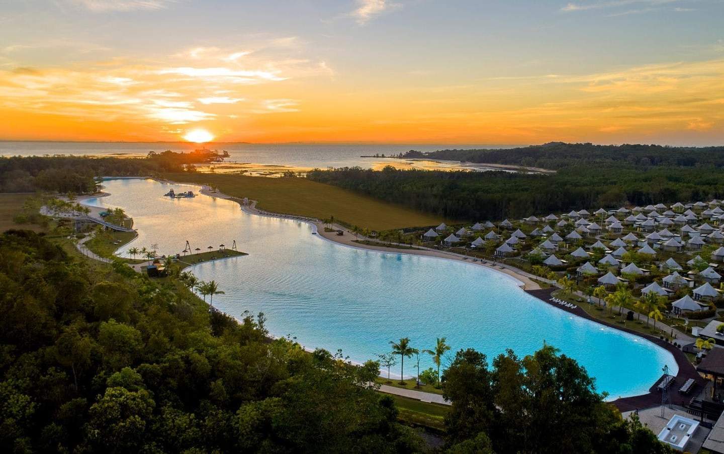 Scenic view of a large lagoon surrounded by greenery, resort huts, and a sunset on the horizon over distant water.