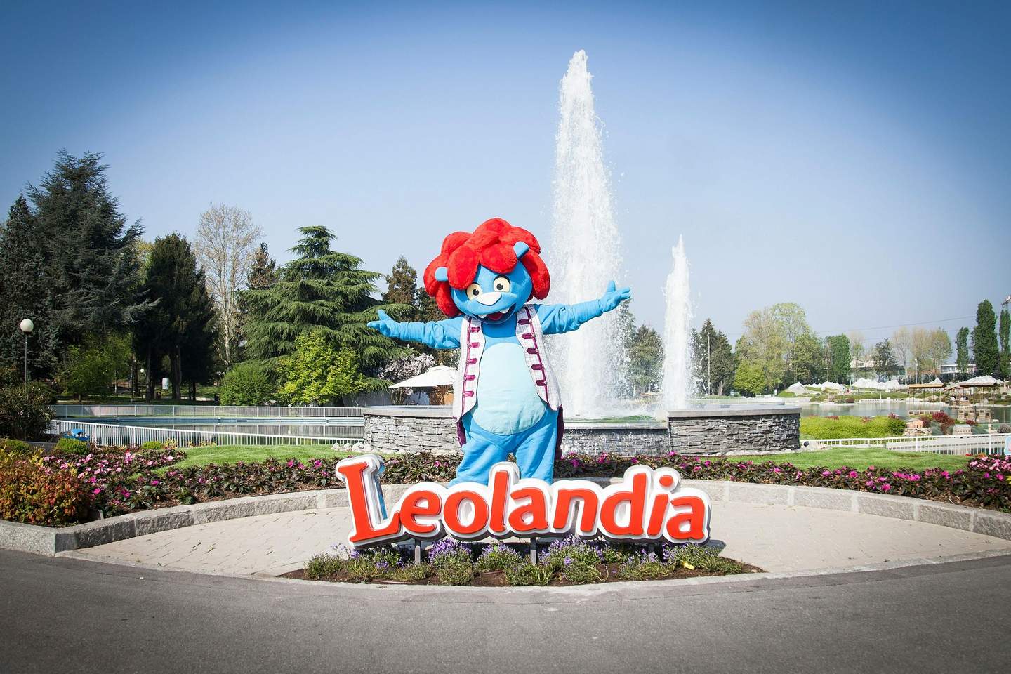 A blue mascot with red hair stands in front of fountains and a "Leolandia" sign in a landscaped park.