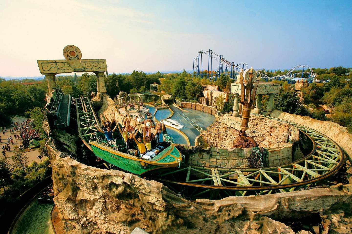 People on a themed roller coaster ride with hands up, surrounded by rocky terrain and ancient structures, with other coasters in the background.