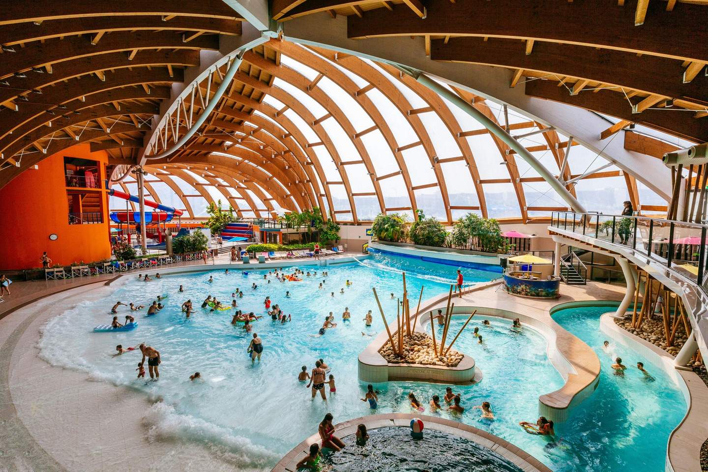 Indoor water park with numerous people swimming in pools, playing in waves, and lounging. Wooden arched roof and water slides.