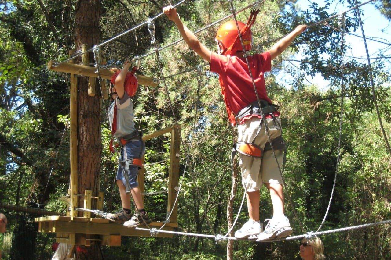 CerviAvventura Two children wearing helmets and harnesses navigate a rope course in a forest setting.