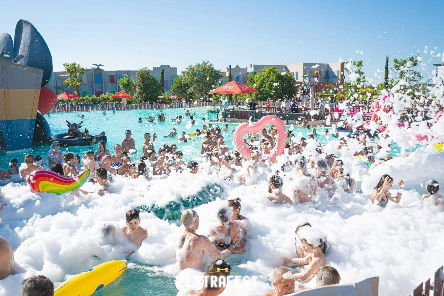 Eine belebte Poolparty mit Menschen in Badekleidung; Schaumblasen, Hüpfburgen und Sonnenschirme sind unter einem klaren blauen Himmel zu sehen.
