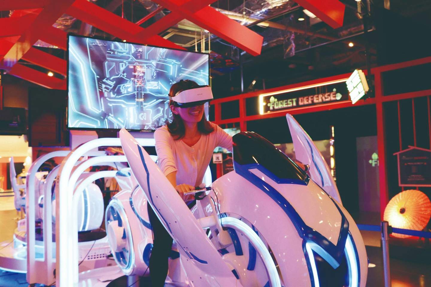 A person using a virtual reality headset and riding a futuristic motorcycle simulator in an arcade setting.