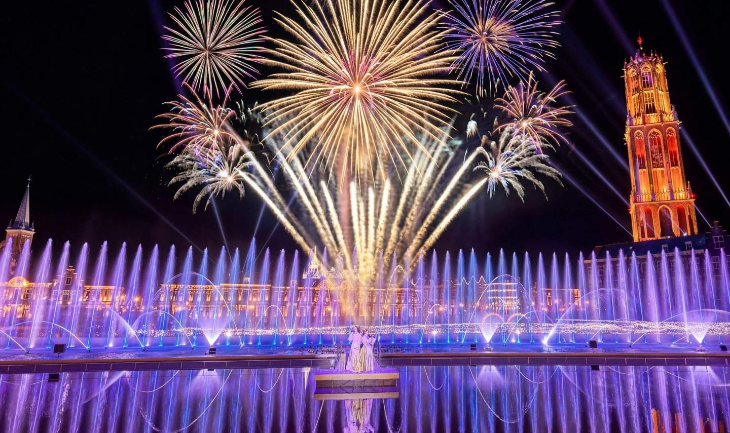 Bright fireworks illuminate the night sky above a lit water fountain display, creating a vibrant and festive scene.