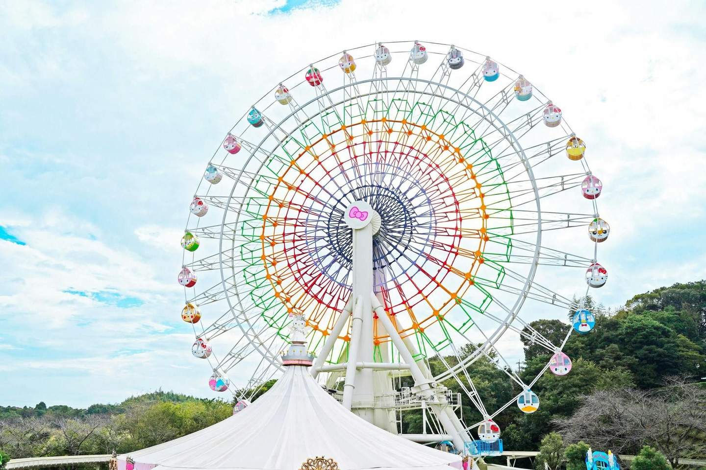 A colorful ferris wheel with themed gondolas against a partly cloudy sky, with trees and a white tent in the foreground.