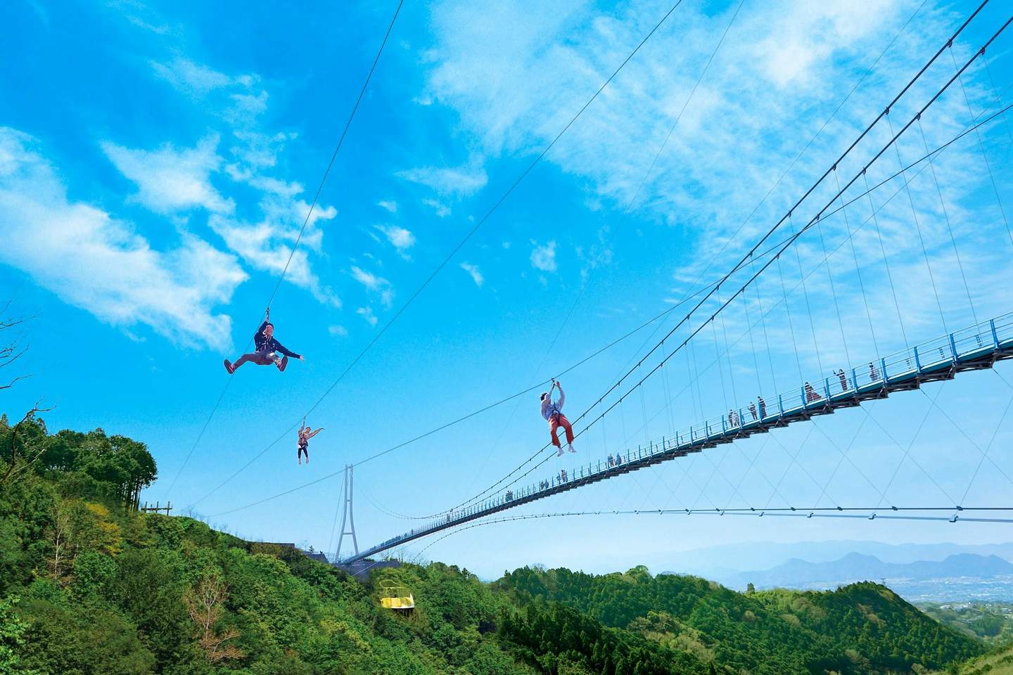 Des personnes font de la tyrolienne au-dessus d'une forêt verdoyante, près d'un pont suspendu, sous un ciel d'un bleu éclatant parsemé de nuages.
