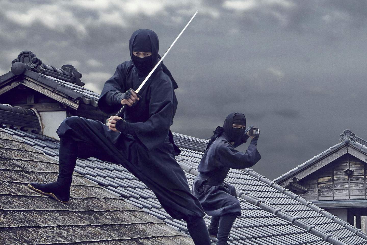 Two ninjas in black attire on tiled rooftops, one holding a sword in an action pose, the other looking through binoculars.