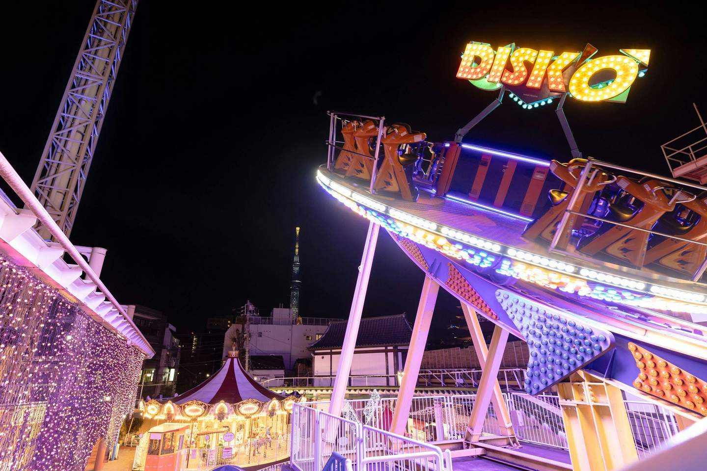 An amusement park ride named 'Disko' illuminated at night, with a carousel and skyscraper in the background.