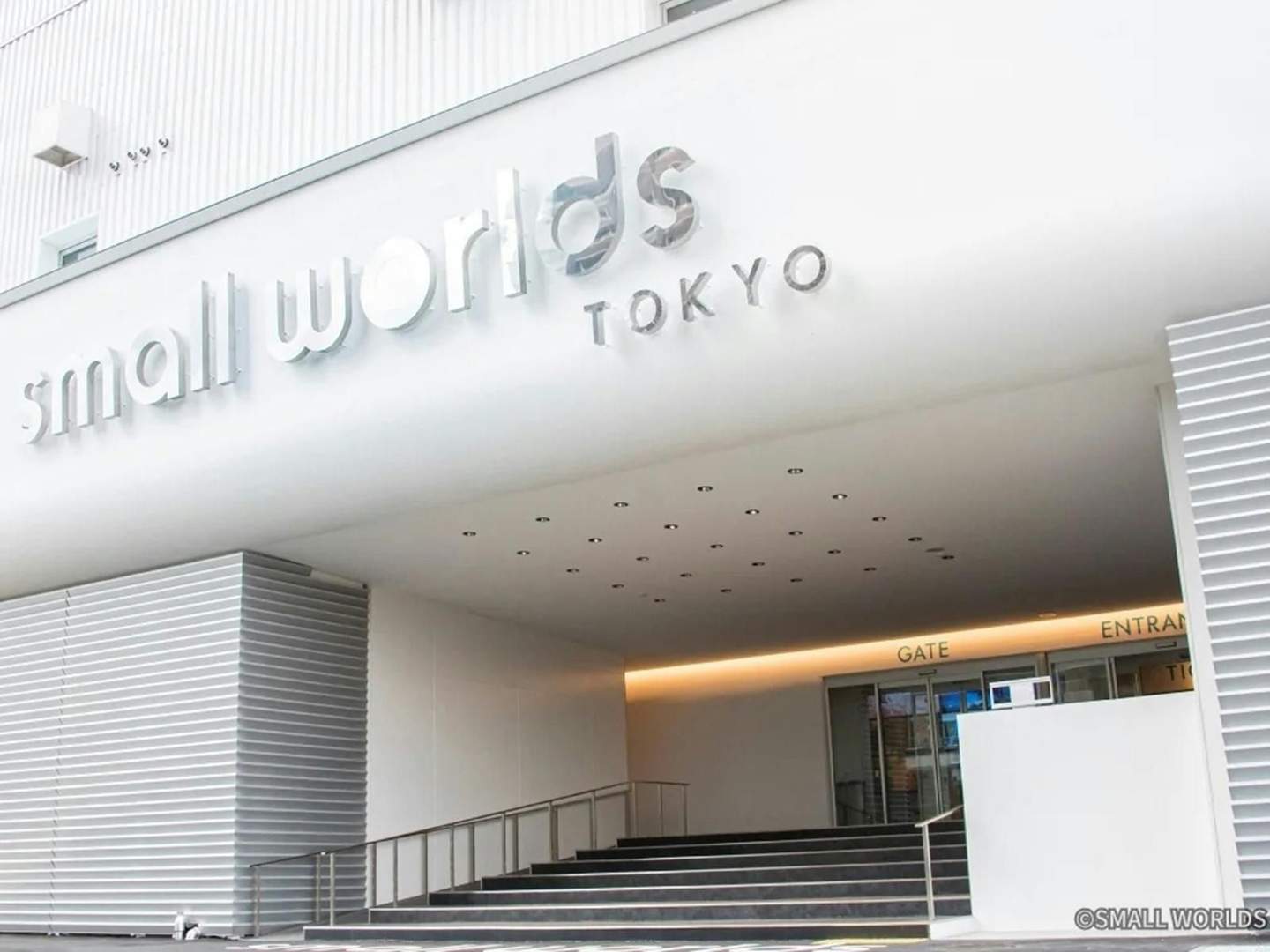 Entrance to Small Worlds Tokyo with a white exterior, metal handrails, and large entrance sign above the stairs and door.