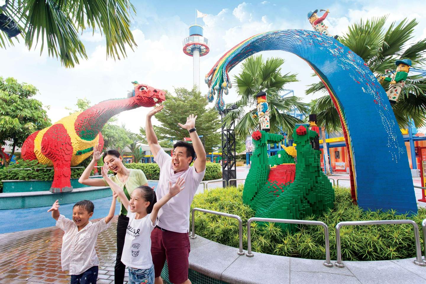 LEGOLAND® Malaysia A family of four cheers beside colorful LEGO sculptures of a dragon, rainbow, and animals at an outdoor amusement park.