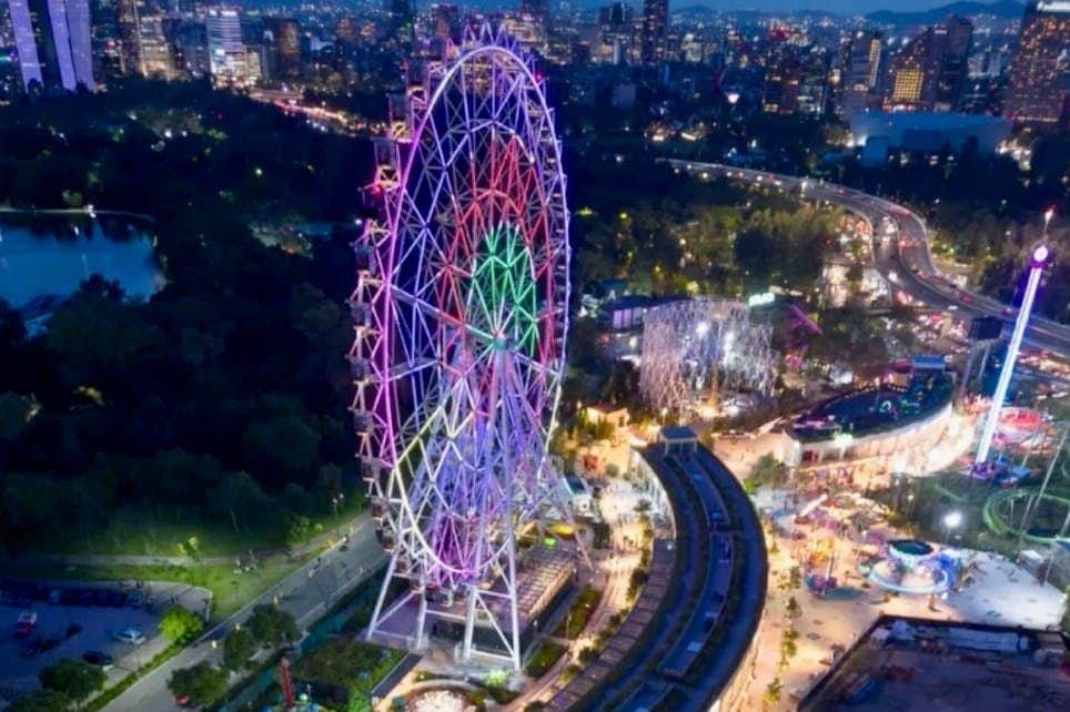 Illuminated Ferris wheel in amusement park at night, with city lights and roadways in the background.
