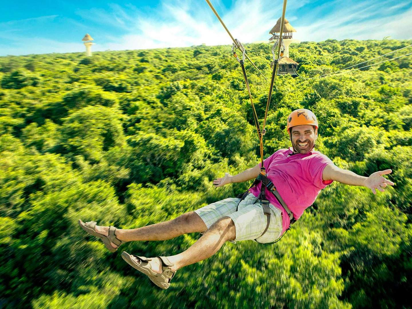 A man in a pink shirt and helmet is zip-lining over a dense forest, smiling with arms outstretched. A few towers are visible in the background.