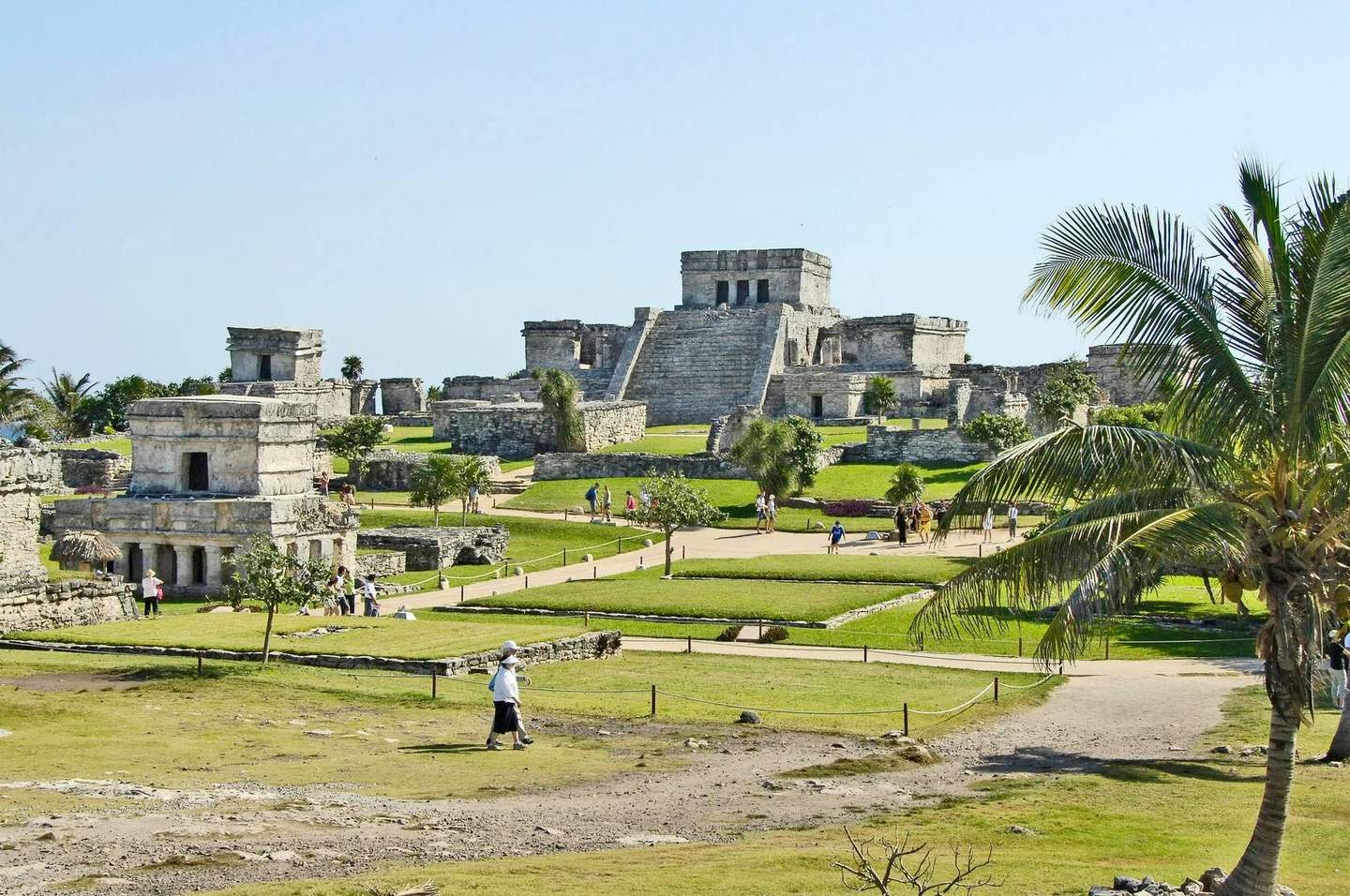 Ancient stone ruins with pyramids and temples, visitors walking among the structures, and a palm tree in the foreground.