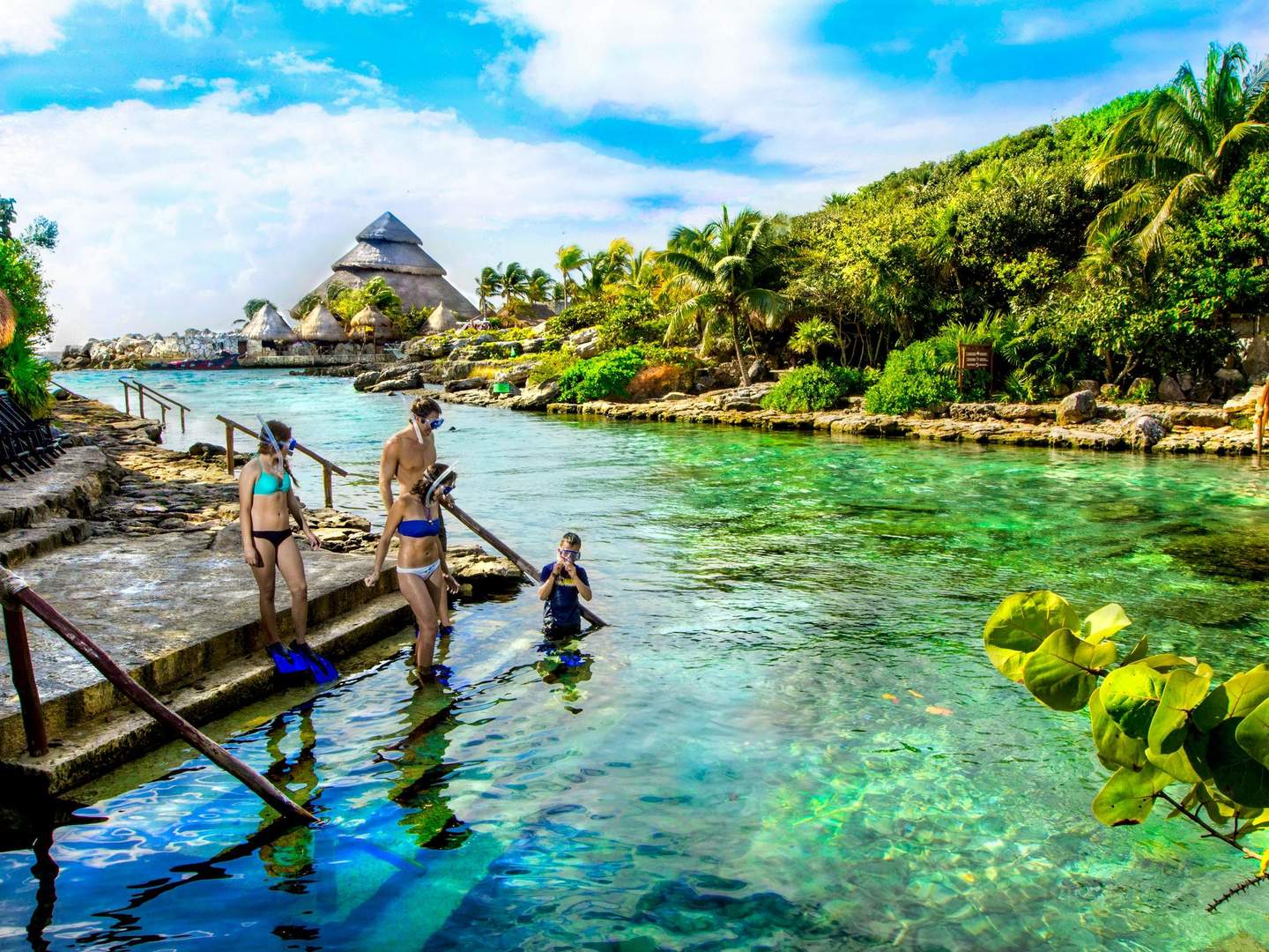 Pessoas em fatos de banho e snorkels preparam-se para entrar numa lagoa tropical límpida, rodeada por uma vegetação luxuriante e estruturas com telhados de colmo.