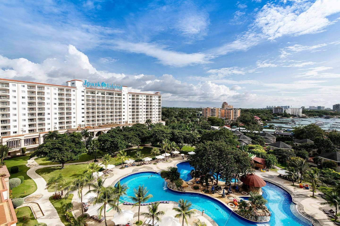 A resort with a large hotel building, landscaped gardens, and a pool area under a clear blue sky.