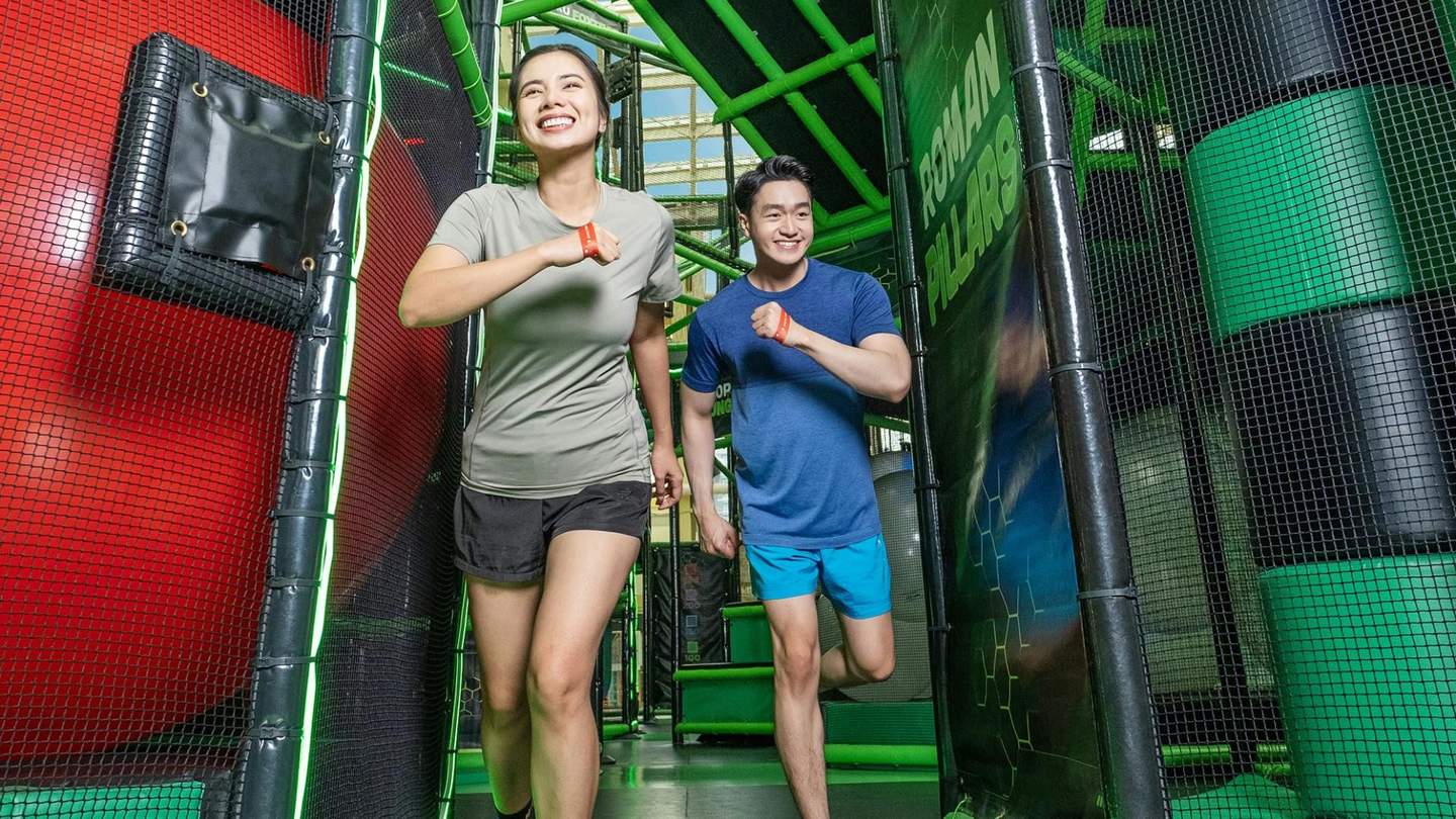Deux personnes qui courent en souriant à l'intérieur d'un parcours d'obstacles couvert, composé de structures vertes et noires.