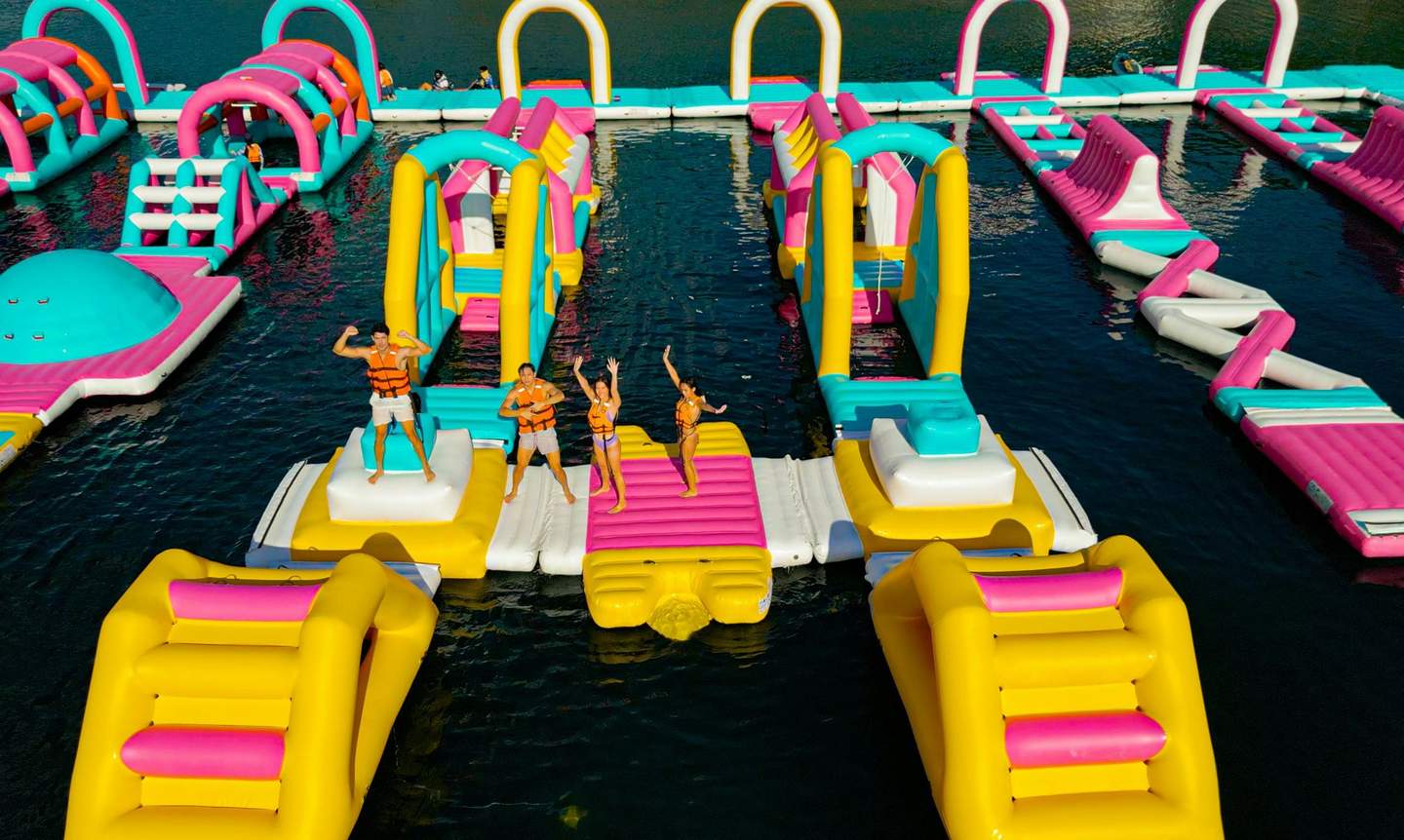 People enjoying a colorful inflatable water park with slides, paths, and structures on a calm body of water.