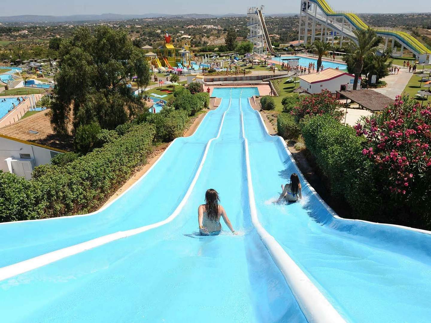 Two people slide down side-by-side water slides in a water park with pools, greenery, and other attractions visible in the background.