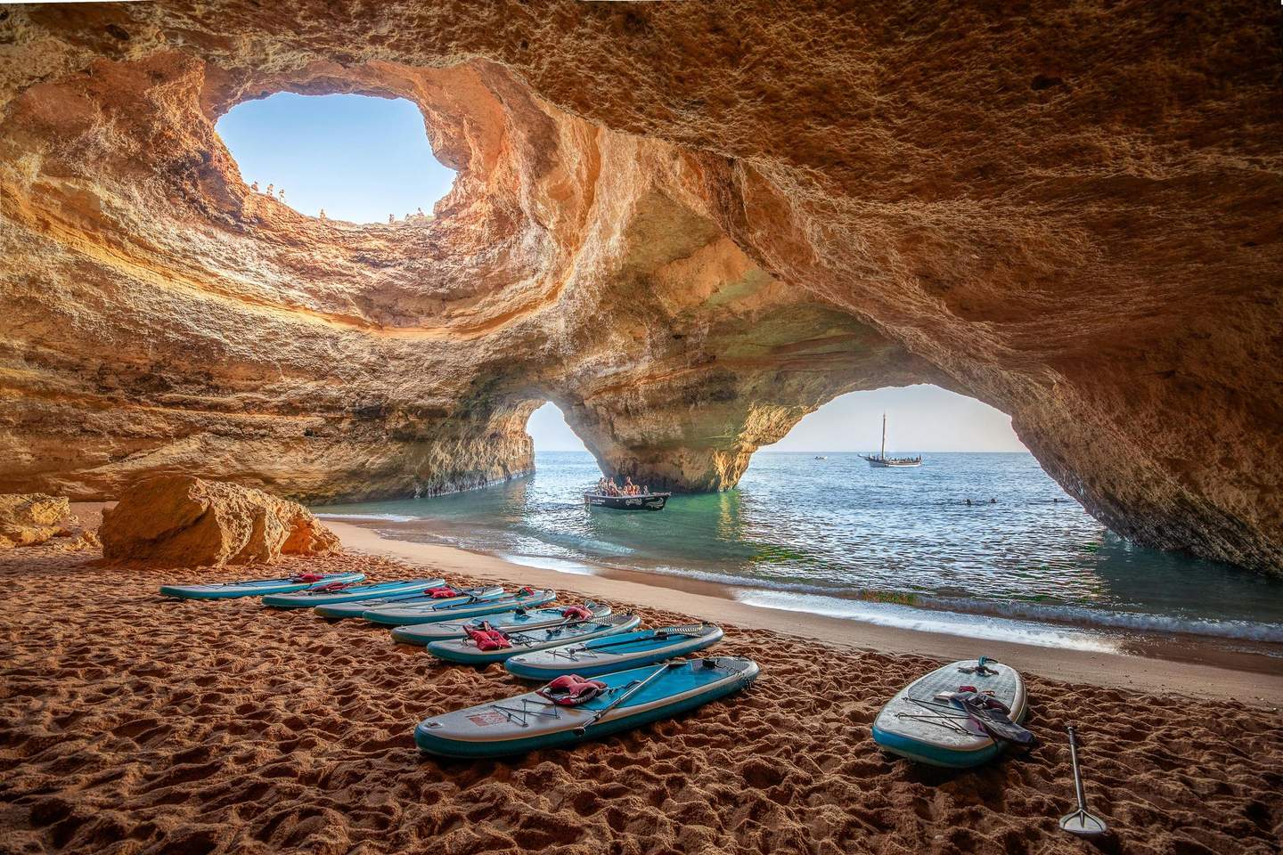 Paddleboards lie on a sandy beach inside a cave with openings revealing boats on blue water.