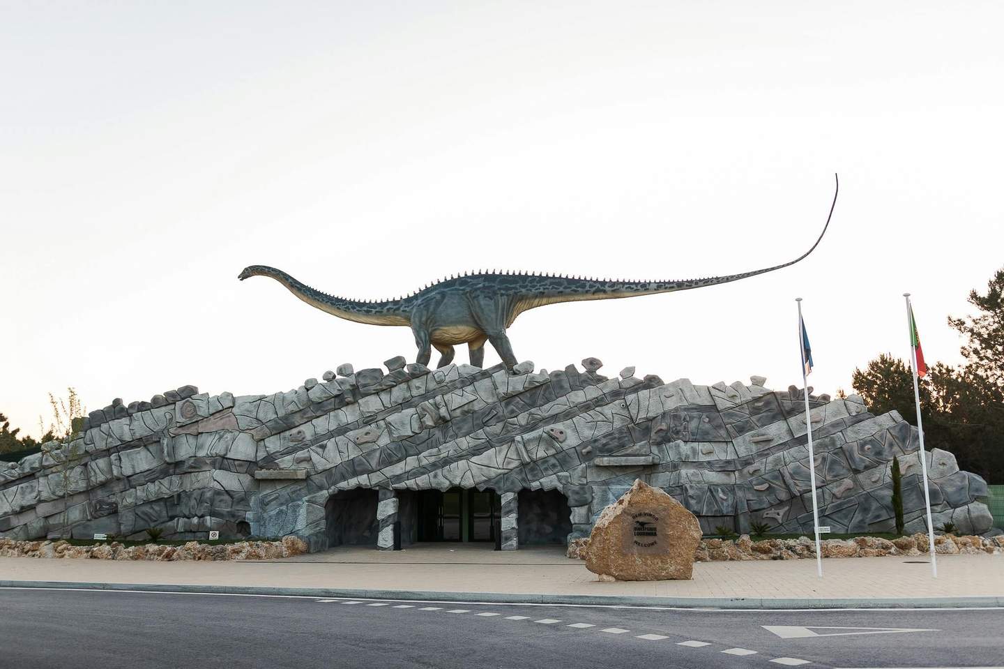 A large dinosaur statue stands atop a building with a rocky exterior. Three flags are placed in front, next to a sign on a stone block.