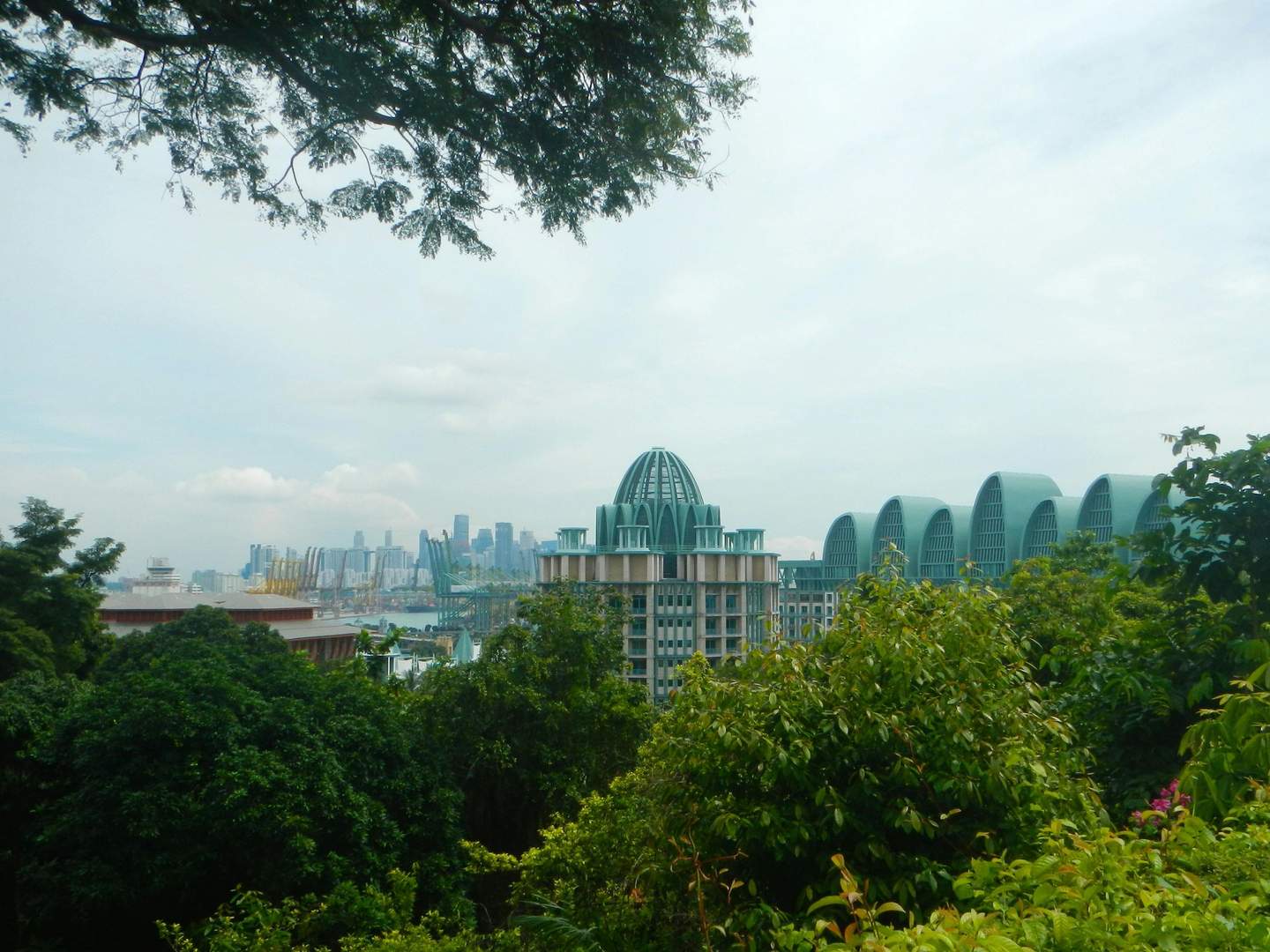 Modern city skyline with unique green-roofed buildings surrounded by trees, under a cloudy blue sky.