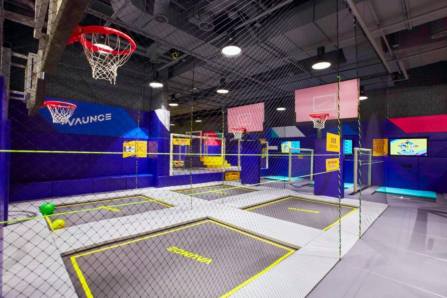 Parque de camas elásticas Vaunce Parque de camas elásticas cubierto con canastas de baloncesto, paredes de colores vivos y redes de seguridad. La marca «Vaunce» se ve en las camas elásticas.