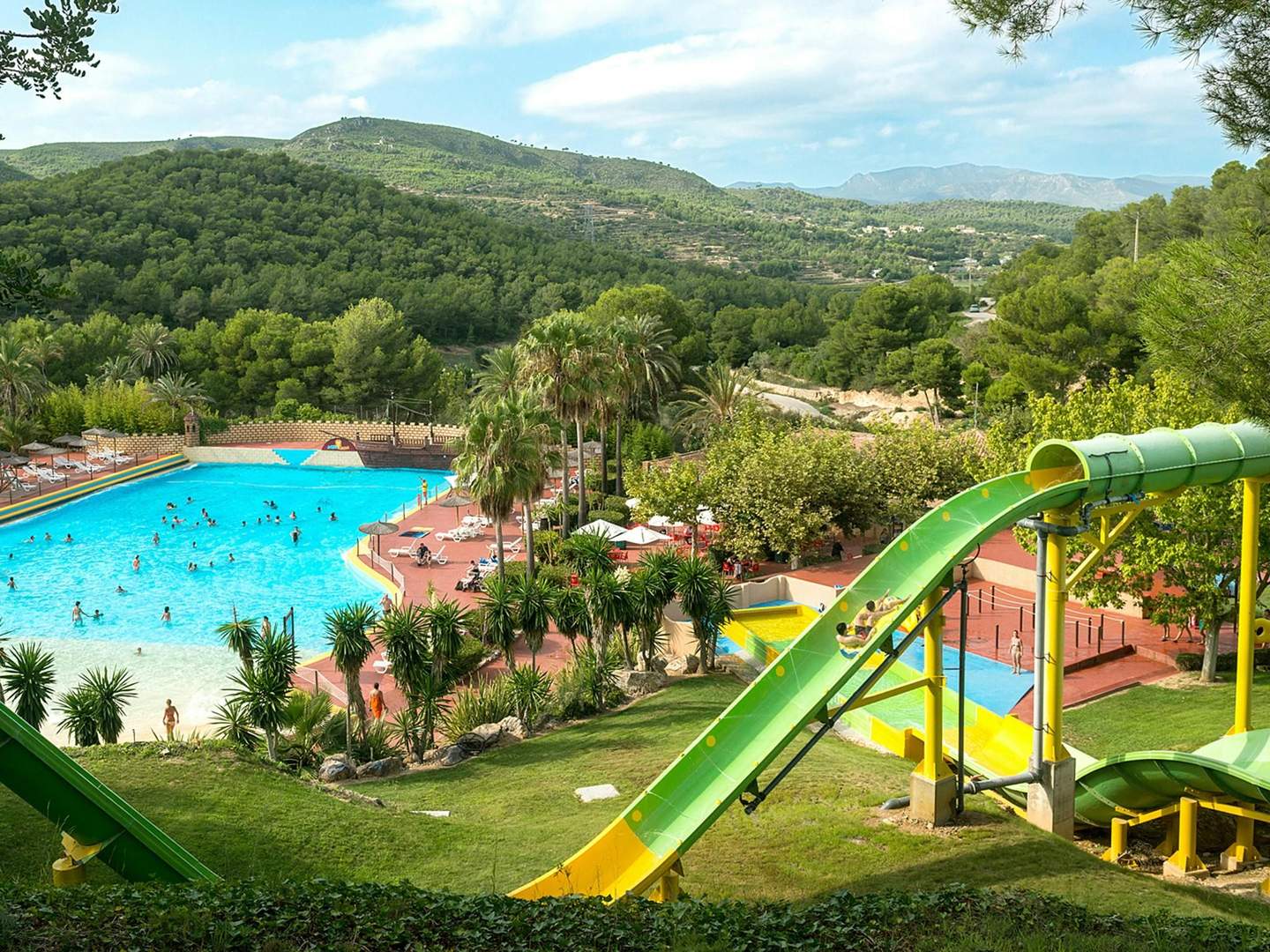 Aqualeon Water Park People enjoying a large outdoor swimming pool and water slides surrounded by lush greenery, hills, and palm trees.