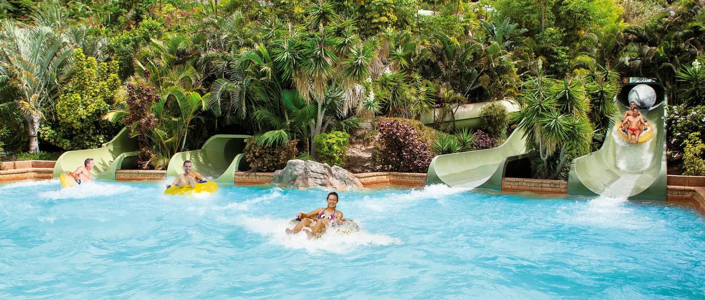 People enjoying water slides into a pool surrounded by lush greenery and tropical plants.