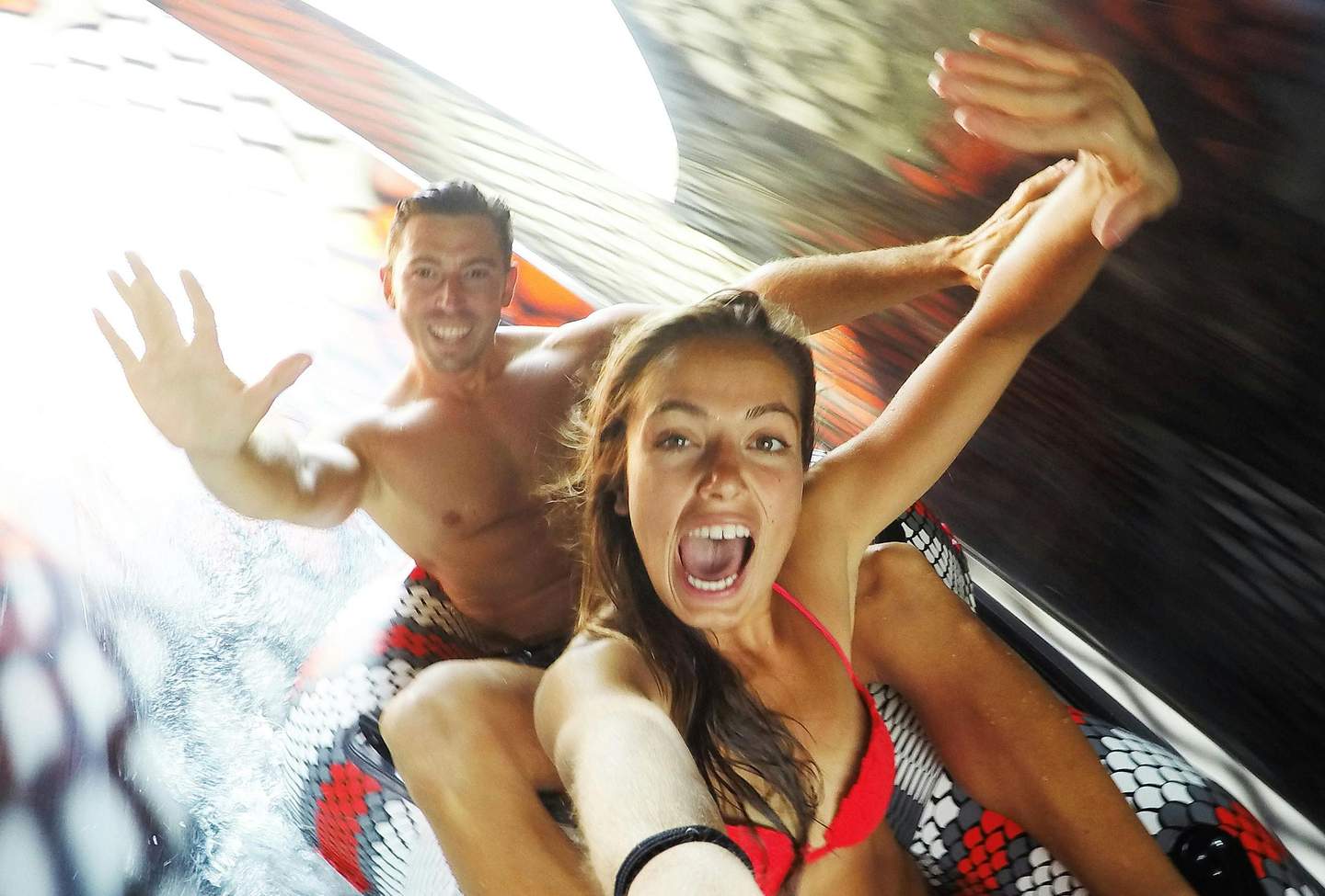 Two people in swimwear are riding an inflatable raft on a water slide, expressing excitement with wide smiles and raised arms.