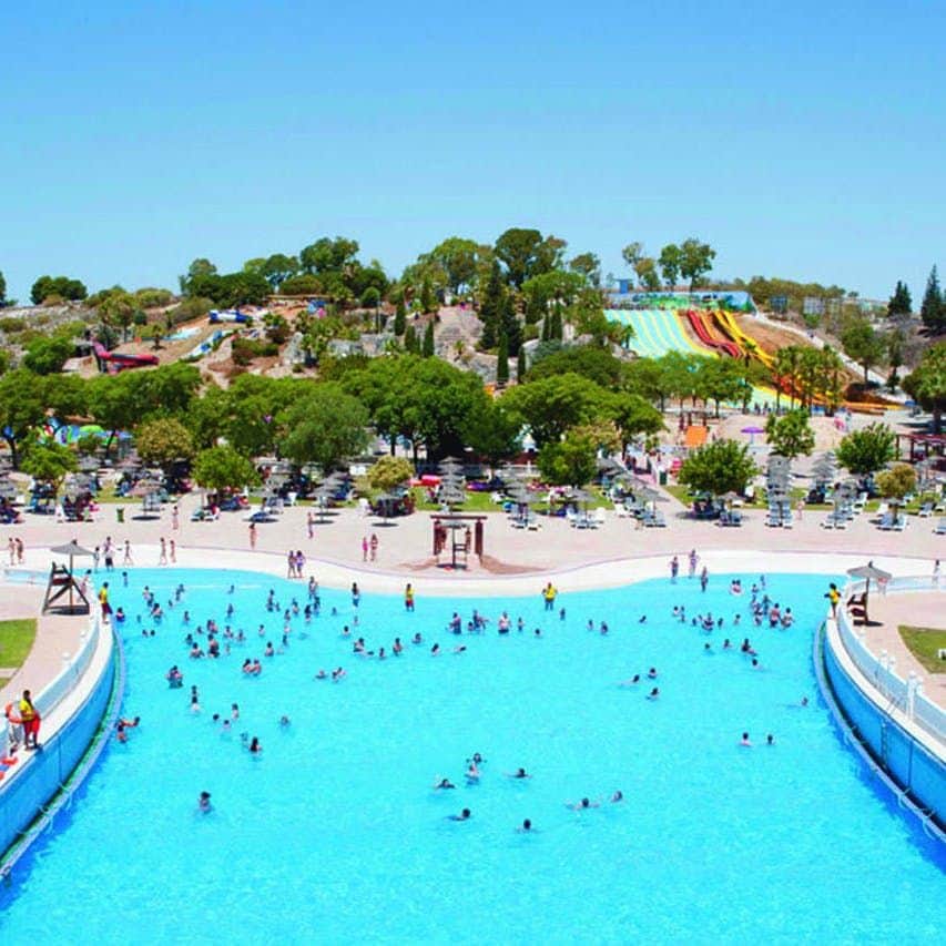 People enjoying a large wave pool at an outdoor water park with colorful water slides and trees in the background.