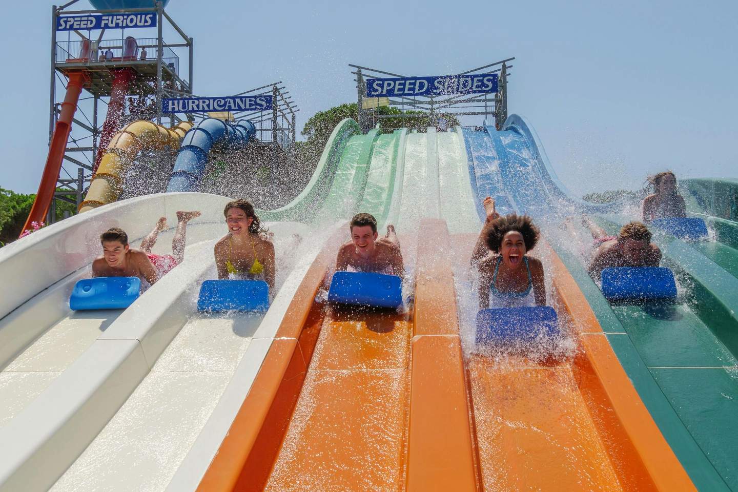 People on colorful water slides, sliding down with excitement, creating splashes, under a clear blue sky.
