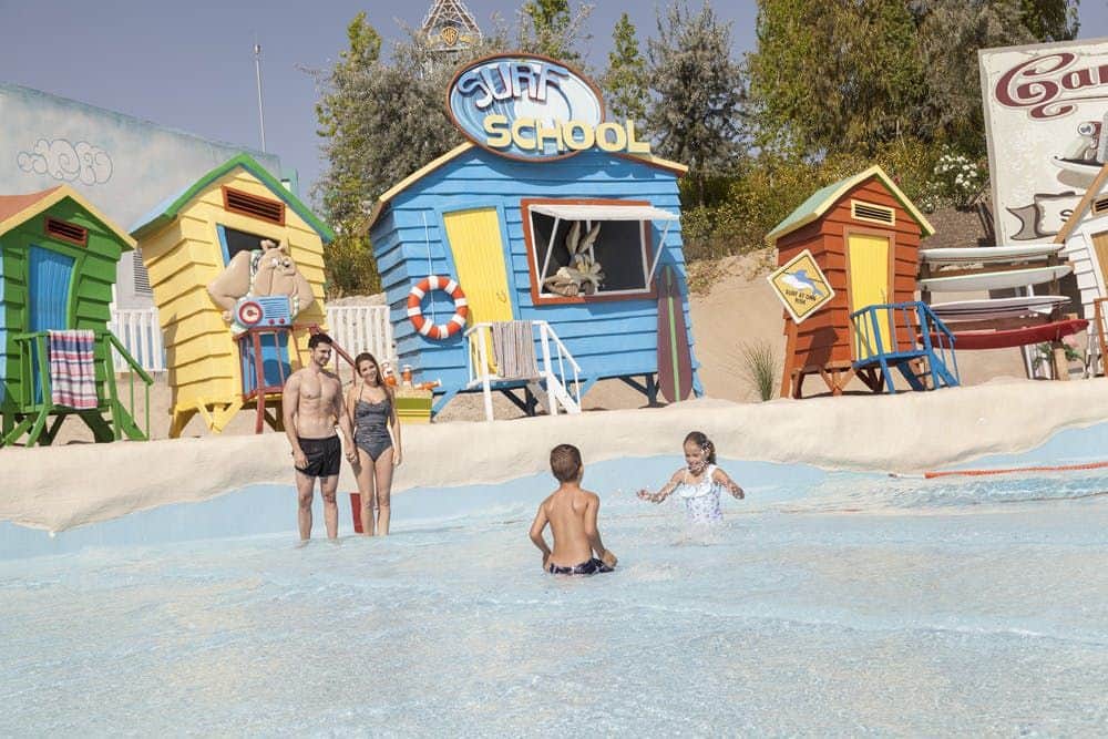 Two children play in a shallow pool, while two adults stand nearby. Colorful beach huts with "Surf School" sign in the background.