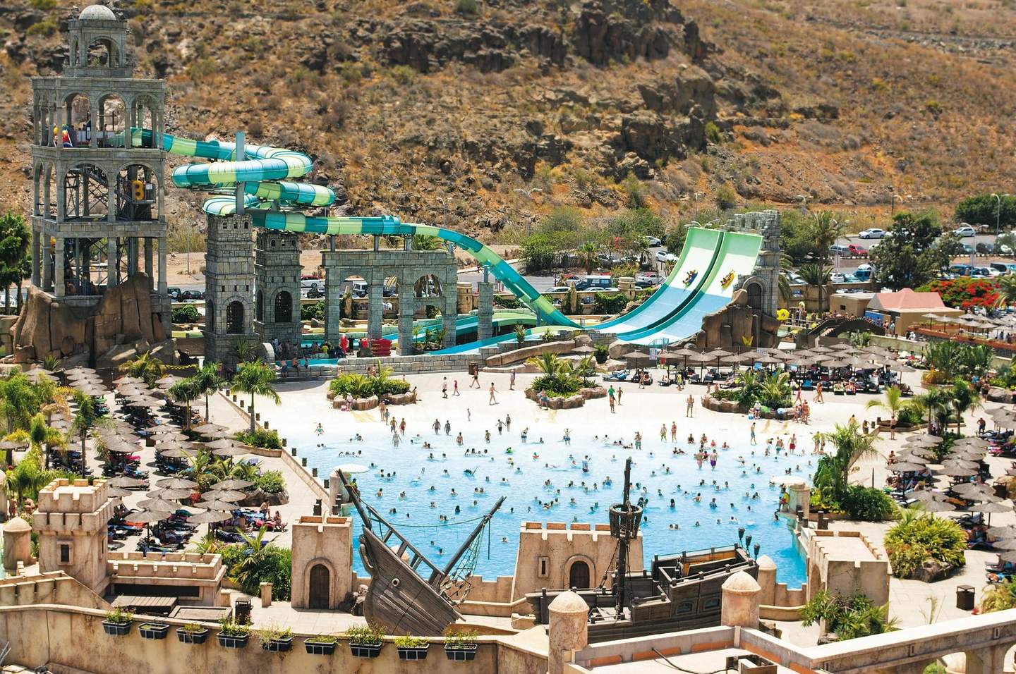 A vibrant water park with people in a wave pool, surrounded by water slides, sun loungers, palm trees, and stone-themed architecture.