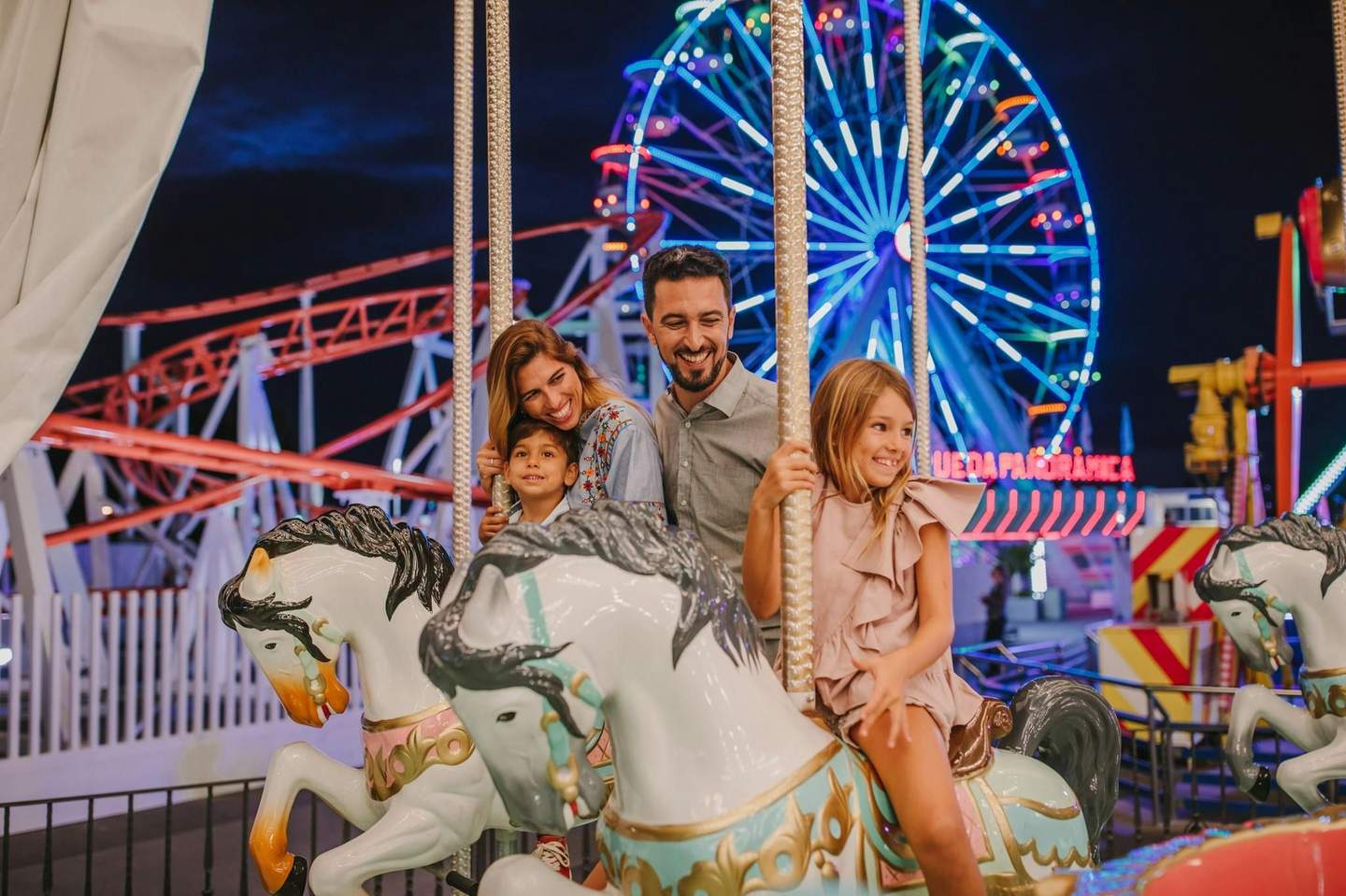 A happy family rides a carousel at night with a lit Ferris wheel and roller coaster in the background.