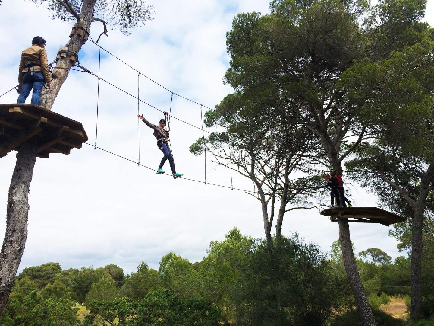Tirolinas Go Mallorca Forestal Park A person in a harness walks across a suspended rope bridge between trees, with two others standing on platforms at opposite ends.