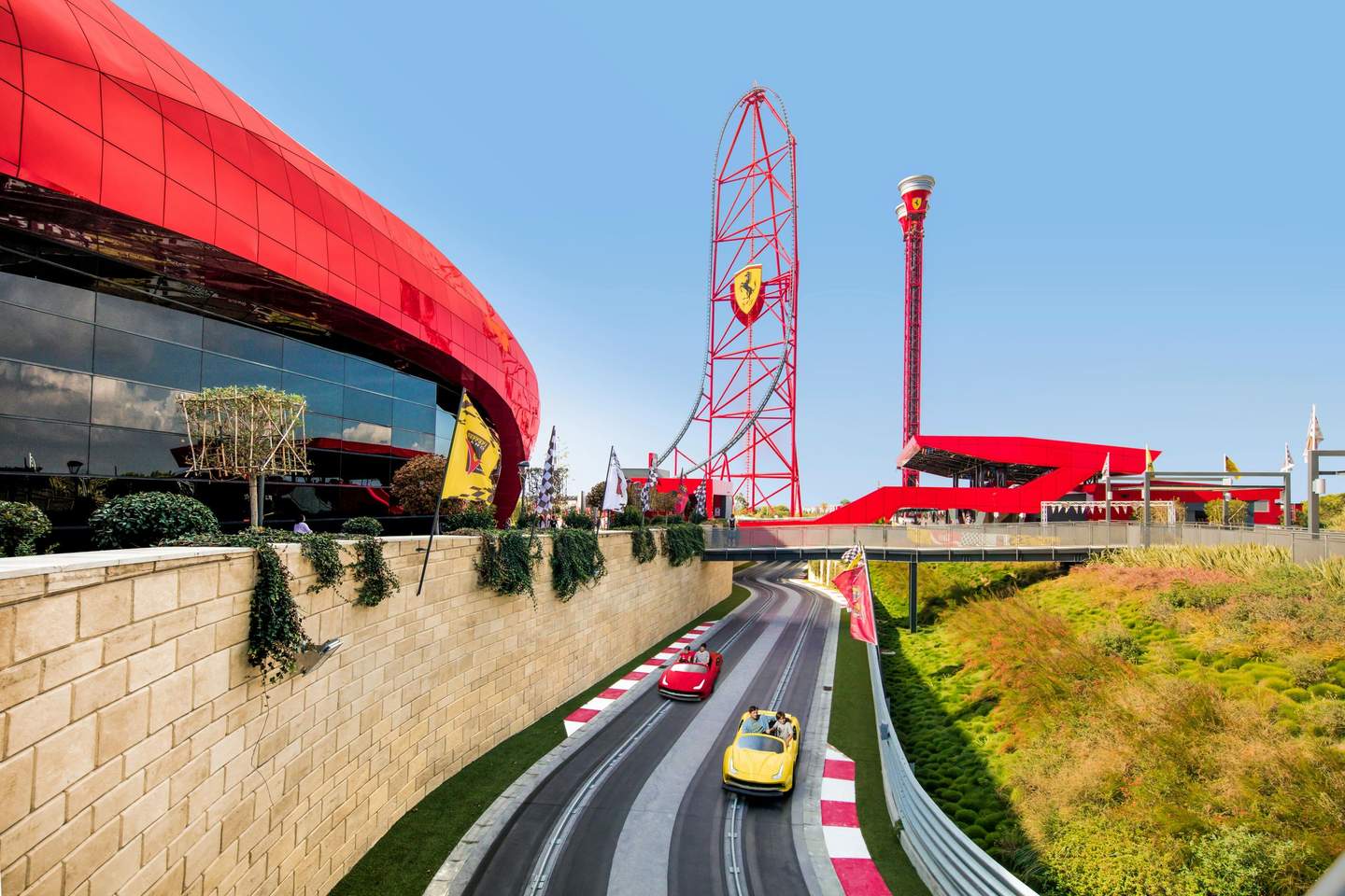 People in red and yellow cars on a track near a red building and tall red amusement park structures against a clear blue sky.