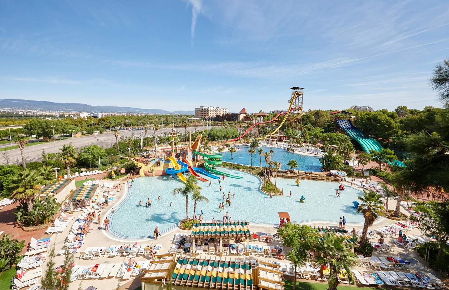 A vibrant water park with various slides, pools, lounge chairs, and palm trees. People are swimming and enjoying the attractions.