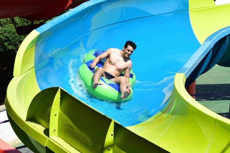Illa Fantasia Waterpark A man rides a green and blue inflatable tube down a brightly colored water slide on a sunny day.