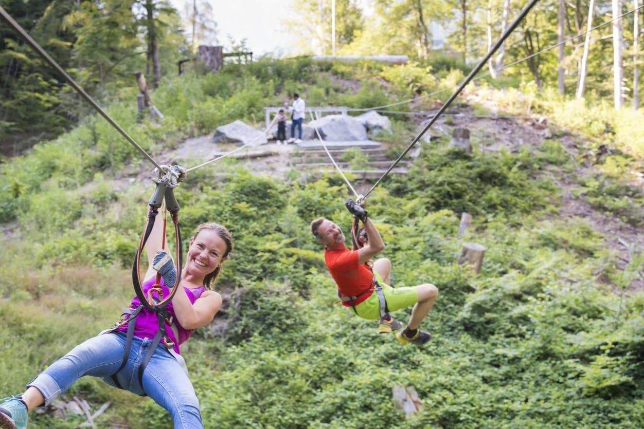 Ropes Park Interlaken Two people smiling while zip-lining through a lush, green forest; two observers are in the background standing on a pathway.