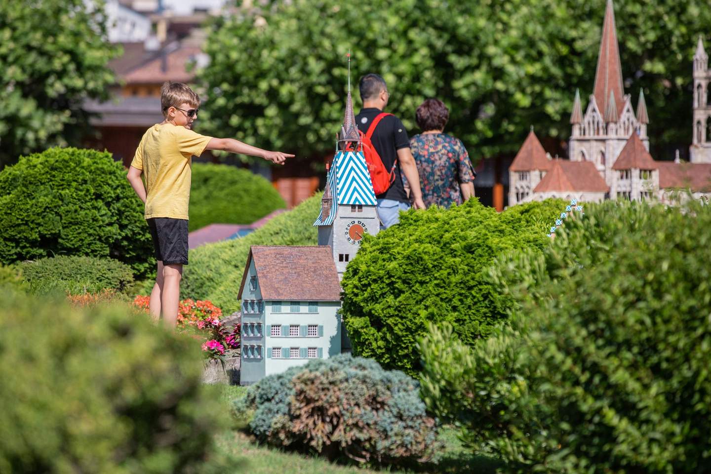 A boy points at miniature buildings in a park while two adults walk nearby, surrounded by lush greenery.