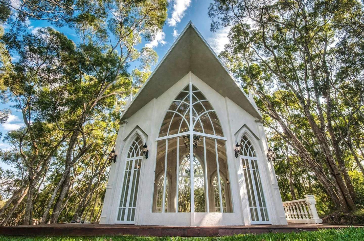A small white chapel with arched windows surrounded by tall trees under a blue sky with scattered clouds.