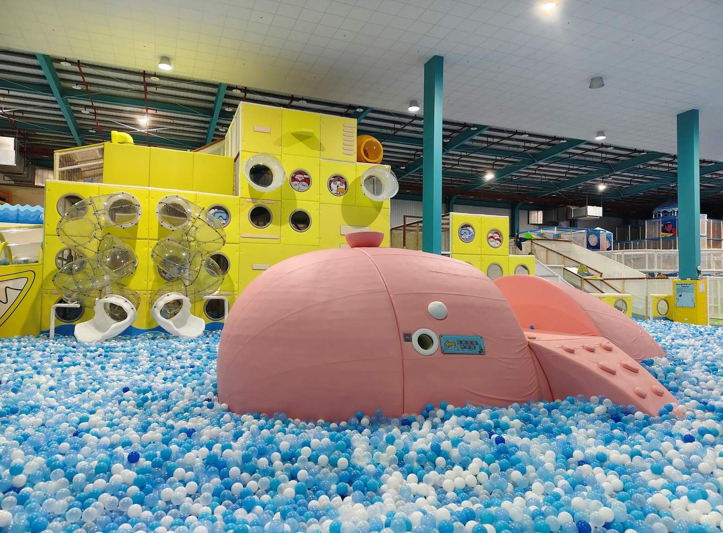 Indoor play area with a pink whale structure in a ball pit and yellow climbing walls with tunnels and slides.
